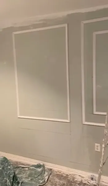 Wall during work, with new white picture-frame molding installed over gray paint and drop cloths on the floor