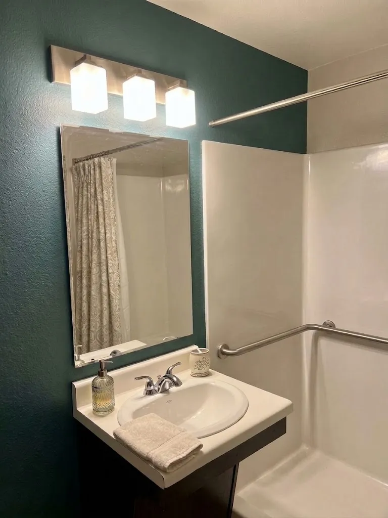 Finished rental bathroom with a dark teal accent wall, a three-light vanity fixture, a white tub-shower surround and a grab bar