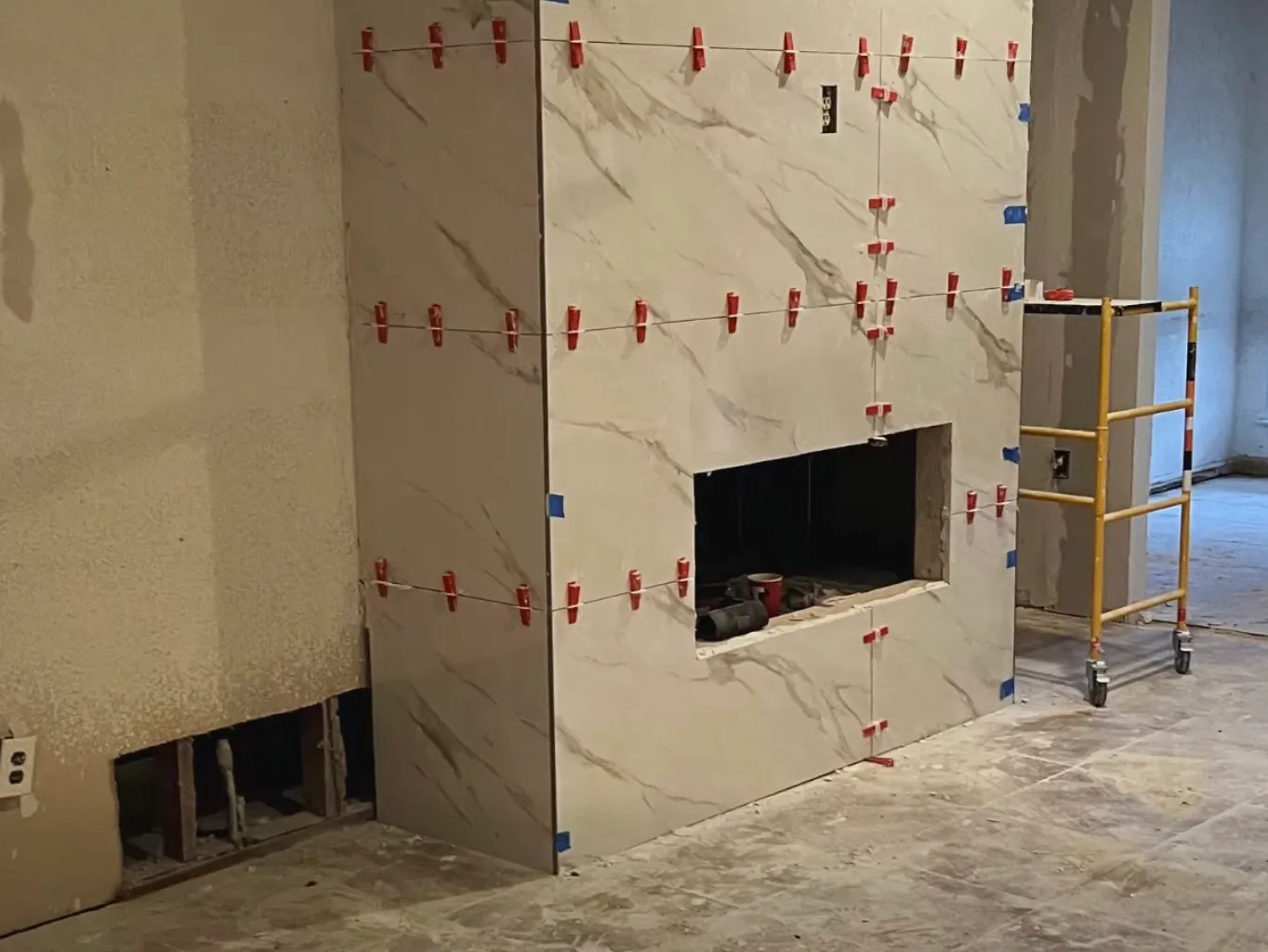 Fireplace bump-out during tiling, with large marble-look tiles held by red leveling clips around the fireplace opening