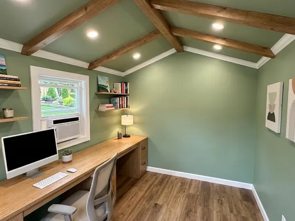 Finished shed office with sage green walls, wood ceiling beams, a built-in wood desk and floating shelves