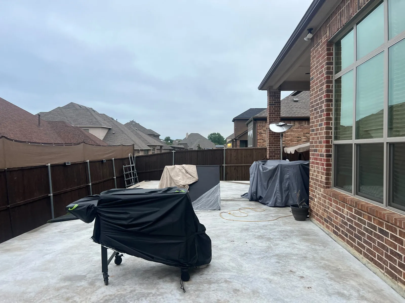 Open concrete patio behind a brick home, with covered grills and a wood privacy fence