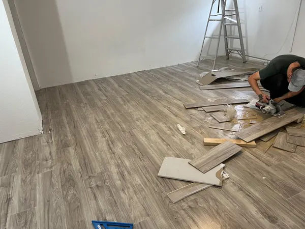 Gray wood-look planks being cut and laid across a room, with a ladder and offcuts on the floor