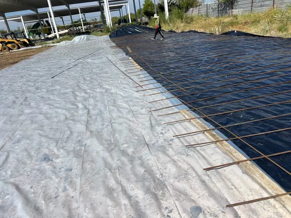 Site prep with plastic vapor barrier sheeting and steel rebar laid out on the ground, a skid steer in the background